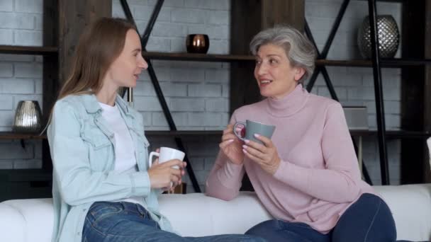 Dojrzała matka i jej dorosła córka piją kawę, rozmawiają i uśmiechają się siedząc na kanapie w domu — Wideo stockowe