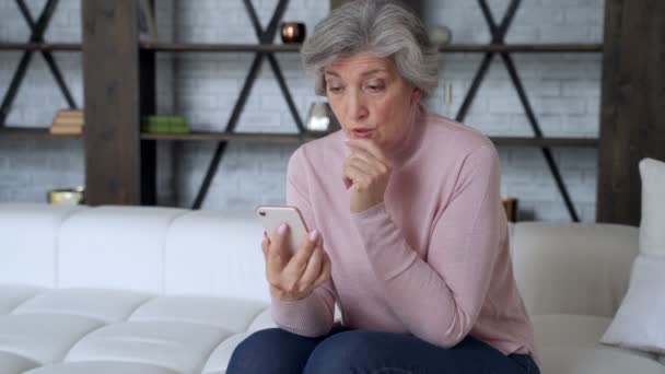Грустная пожилая женщина сидит на диване держать смартфон чувствует разочарование от полученных смс плохие новости, трудности с современным использованием устройства. — стоковое видео
