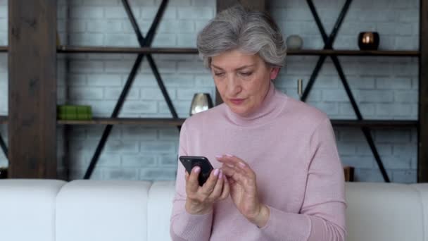 Fröhliche reife Frau sitzt auf dem Sofa und benutzt ein Mobiltelefon — Stockvideo