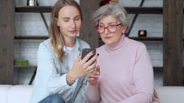 Starsza matka i dorosła córka siedzą na kanapie w domu, bawią się razem za pomocą smartfona, oglądają filmy na telefonie komórkowym. — Wideo stockowe