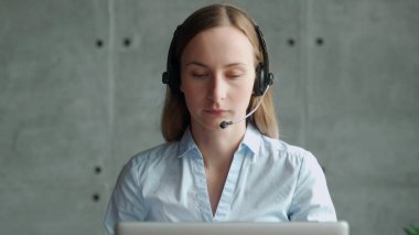 Kablosuz kulaklık takan mutlu bayan yönetici, dizüstü bilgisayara bakıyor, ortaklarla online hoş sohbetler yapıyor, iş yerinde uzaktan çalışıyor.