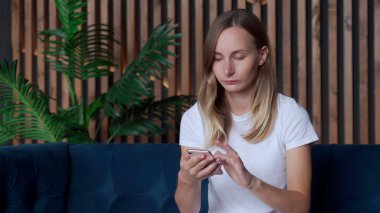 Mutlu kadın rahat koltukta dinleniyor, akıllı telefonu elinde tutuyor. Gülümseyen kadın sosyal ağlarda sohbet ediyor, evdeki mobil uygulamaları kullanıyor..