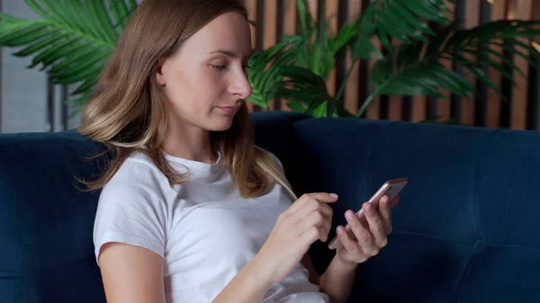 Mujer feliz relajándose en un cómodo sofá, sosteniendo el teléfono inteligente en las manos. Mujer sonriente charlando en las redes sociales, utilizando aplicaciones móviles en el hogar. — Foto de Stock