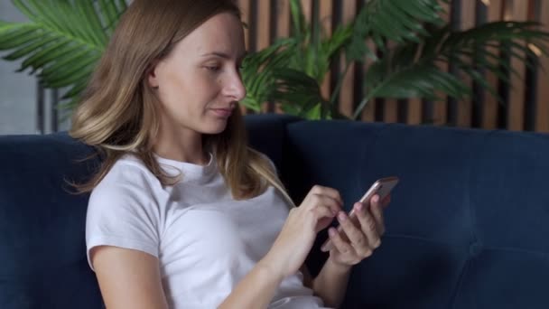 Gelukkige vrouw ontspannen op de comfortabele bank, het houden van smartphone in handen. Lachende vrouw chatten in sociale netwerken, met behulp van mobiele toepassingen thuis. — Stockvideo