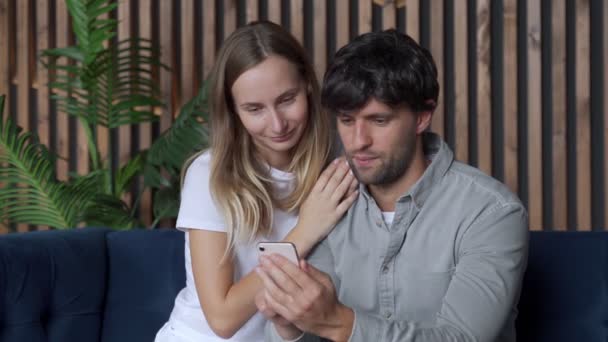 Sorrindo jovem casal usando smartphone juntos no sofá em casa — Vídeo de Stock