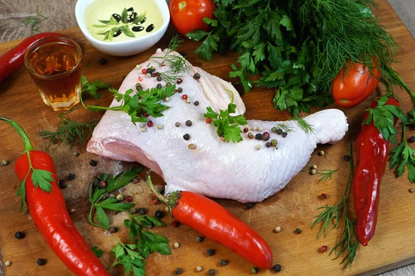 A quarter of the chicken is raw meat on a wooden board next to herbs and chili peppers, oil and vinegar.     selective focus