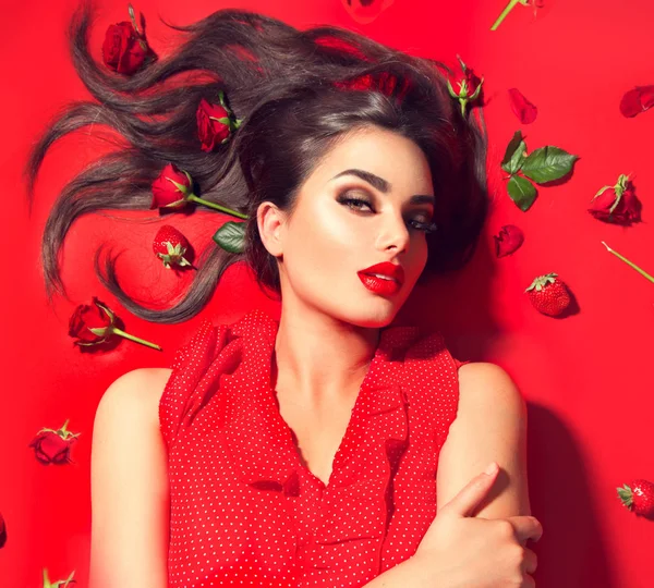 バラの花とイチゴと赤い背景に横たわっている美しさセクシーなモデルの女の子 長い髪と完璧なメイクアップ 赤い魅惑的な唇 スモーキーな目を持つ美しいブルネットの若い女性 トレンディなメイクアップ ロイヤリティフリーのストック画像