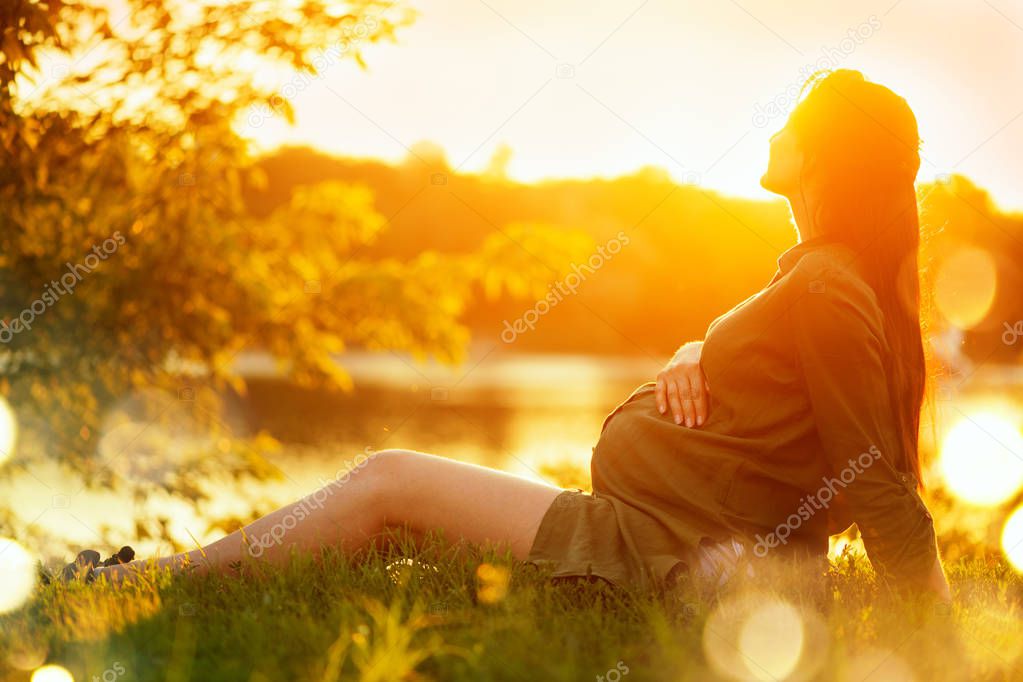 Pregnant woman sitting on green grass in summer park, enjoying n