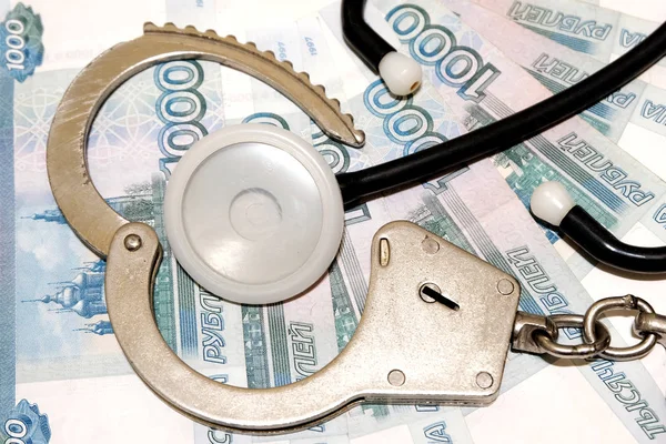 Tıp alanında hukukun ihlali. Polis kelepçe açık bir bilezik ve tıbbi phonendoscope Rus parası arka plan üzerinde yalan. Yolsuzluk Rusya'da hekimlerin.