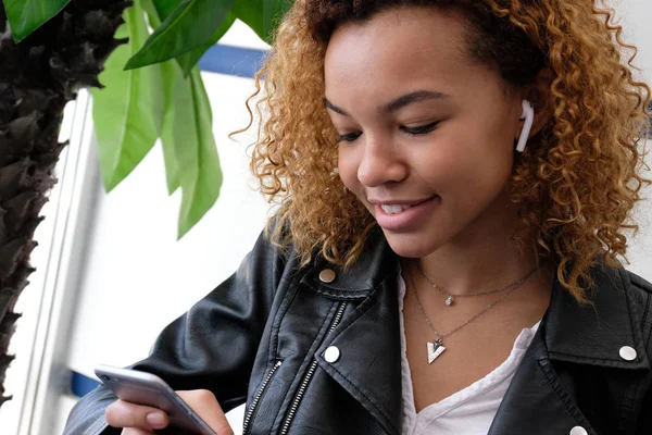 Afro-Amerikan Kız gülümseyerek, telefonunu arıyorum. Güzel genç modern bir siyah kadın, onun kulağına airpods ile deri ceketli müzik dinliyor.
