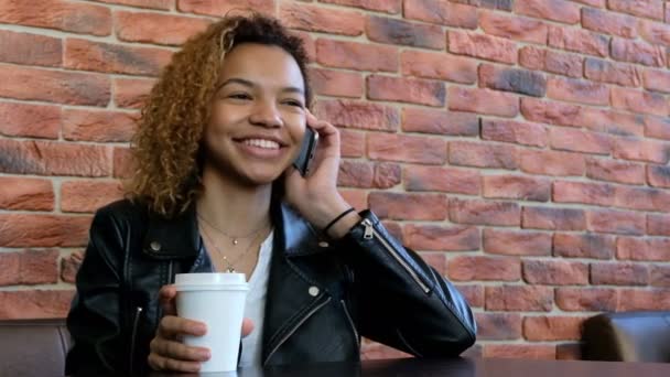 Uma jovem e moderna menina afro-americana está sorrindo falando ao telefone e bebendo uma bebida de uma xícara branca. A nitidez e o foco se movem de pessoa para smartphone. Atire o vídeo das mãos . — Vídeo de Stock