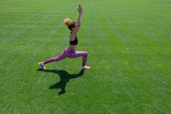 Une jeune afro-américaine vêtue d'un T-shirt noir, d'un pantalon rose et de baskets faisant des exercices sportifs sur de l'herbe verte et levant les mains. Style de vie moderne. Espace de copie . — Photo