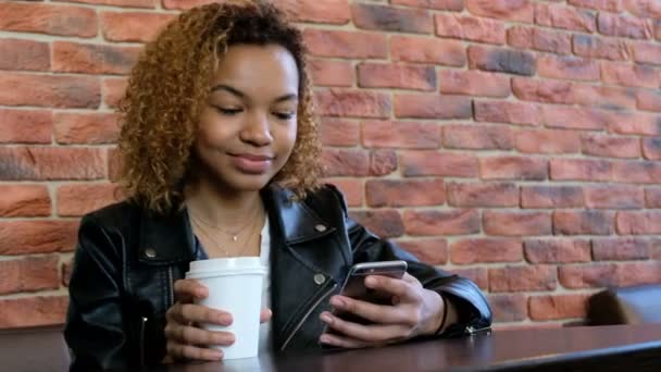 Uma jovem moderna e bela menina afro-americana segurando uma xícara branca e telefone e dizendo algo. Atire o vídeo das mãos . — Vídeo de Stock