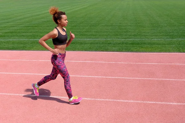 Un afroamericano en los deportes de verano. Una joven sexy y deportiva chica negra corre a lo largo del camino rosa del estadio junto a la hierba verde . — Foto de Stock