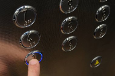 Asansörde erkek el parmağı mavi renkte yanıyor birinci kat düğmeye basmadan. Yakın çekim. Düğmeleri numaraları ile yuvarlak metal yüzey.