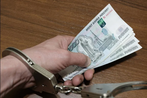 A close-up of a male hand holding a bundle of banknotes of Russian rubles with handcuffs lie on a brown background. Concept of violation of the law in the financial sphere, corruption, bribery, fraud.