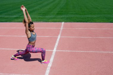 Spor egzersizleri ve stadyumda germe. Genç esmer kız Gri tişört, pembe pantolon ve spor ayakkabı ellerini kaldırdı ve koşu bandı satırın yakınında duruyor. Yan görünümü. Boşaltmak