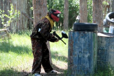 Bir genç kız ya da çocuk benekli koruyucu elbise ve kırmızı bir koruyucu maske takarak bir orman aralığını paintball oynuyor. Varil saklanma koruyucu gibi lekeleri, siyah lastikleri ile.