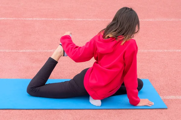 A girl in a red T-shirt and black pants performs fitness exercises. A blue gym mat in the open air. Back view. Training on stretching in a stadium with pink treadmills.