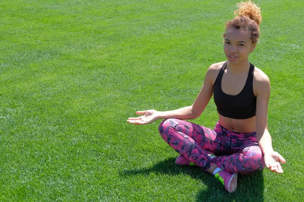 Siyah T-shirt ve pembe ayakkabılarla genç bir Afrikalı-Amerikalı kadın meditatif bir pozisyonda çimenlerin üzerinde oturur ve kameraya benziyor.