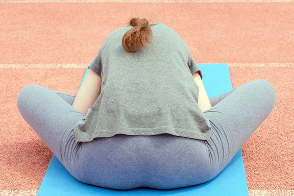 A girl in a gray T-shirt and pants on a blue rug is engaged in sports stretching exercises. Back view. Fitness exercise in a meditative pose at the stadium.