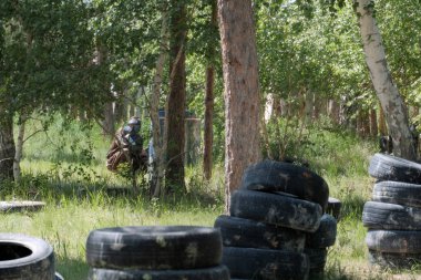Ağaçların arasında çim kolları olan bir siyah koruyucu maske ve kamuflaj kıyafetleri giyen bir genç. Bir takım oyunu Paintball için ormandaki düzenli depolama.