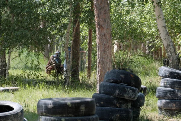 Ağaçların arasında çim kolları olan bir siyah koruyucu maske ve kamuflaj kıyafetleri giyen bir genç. Bir takım oyunu Paintball için ormandaki düzenli depolama.