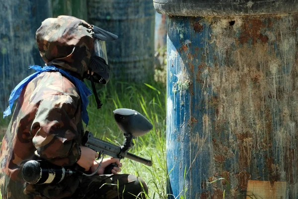 Düşmanın varil arkasında pusuda bekliyor. Maske, silah, kamuflaj ve paintball oyun sırasında oyuncu mavi bir mendil.