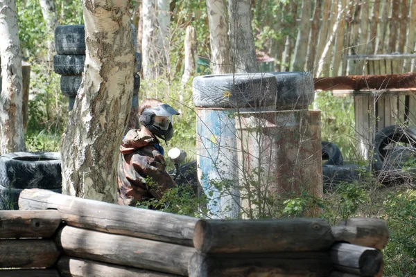 Aşırı takım sporu. Paintball depolama üzerinde bir oyun sırasında bir adam. Kamuflaj takım elbise, koruyucu maske ve silah. Lastikler, varil, orman içinde ahşap çitler.