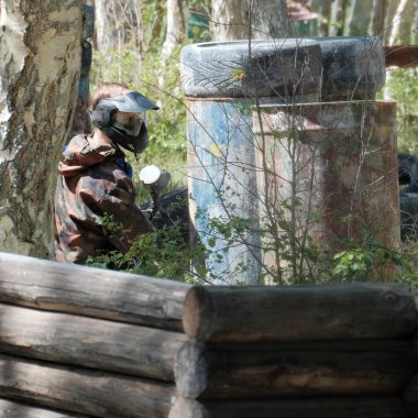Paintball oyuncu saklandığı yerden görünüyor. Kamuflaj takım elbise, koruyucu maske ve silah. Lastikler, varil, orman içinde ahşap çitler. Aşırı takım sporu.