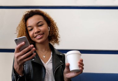 Kablosuz kulaklık kulak bir Afrikalı-Amerikalı genç kadın. Beyaz cam elinde tutan ve seyir telefon mutlu güzel esmer kız.