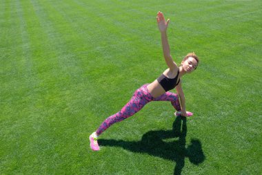 Afrikalı-Amerikalı kız siyah T-shirt, pembe pantolon ve spor egzersizleri yeşil çimenlerin üzerinde yapıyor ve elleri kadar yükselterek spor ayakkabı. Modern spor yaşam tarzı.