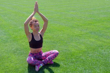 Yaz spor giysiler içinde bir Afro-Amerikan Kız ellerini kaldırdı ve derin derin düşünür. Rahatlatıcı çimenlerin üzerinde oturan genç sevimli siyah kız poz ile kapalı gözler.