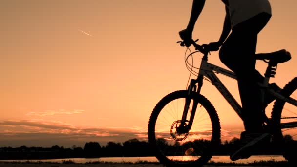 Un adolescente con una maglietta bianca in bicicletta passa davanti a un fiume o a un lago la mattina presto all'alba. Silhouette di un ciclista su uno sfondo di un cielo dorato prima dell'alba. Stile di vita sportivo . — Video Stock