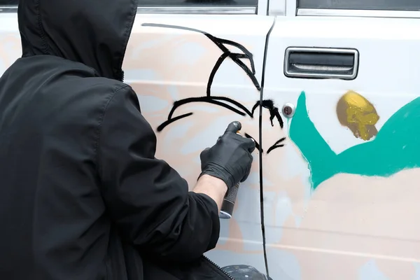 Youth subculture graffiti. A guy in black clothes and gloves draws on a white car an abstract image. Vandalism or art.