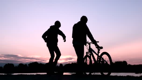 Dois jovens cumprimentam-se e despedem-se. Um adolescente sai de bicicleta e outro num giroscópio de duas rodas. Silhuetas de pessoas ao nascer ou ao pôr-do-sol . — Vídeo de Stock