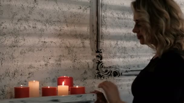 A beautiful blond girl in a black dress approaches the shelf with burning candles. Waiting for an evening meeting or a pre-holiday mood. — Stock Video