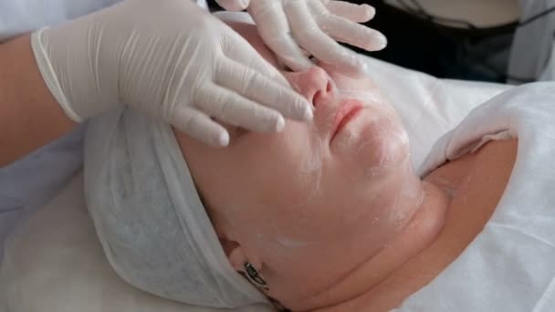 Close-up portret van een dikke vrouw op een procedure voor het reinigen van de huid in een schoonheidssalon. De handen van de schoonheidsspecialist in witte handschoenen wrijf zeep schuim op het gezicht van het meisje in het midden van de cosmetologie. — Stockvideo