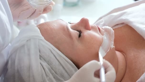 A woman receives a procedure for cleaning the skin in a beauty salon. The hands of the cosmetologist in gloves smear with a brush a white cream on the face of a fat girl in a cosmetology center. — Stock Video