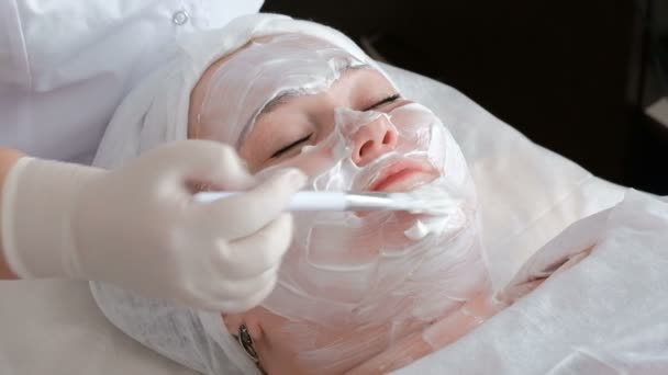 一个胖女人在美容院办公室接受清洁和解除脸部皮肤的程序。美容师的手用刷子涂抹在一个女孩的额头上的白色奶油 — 图库视频影像