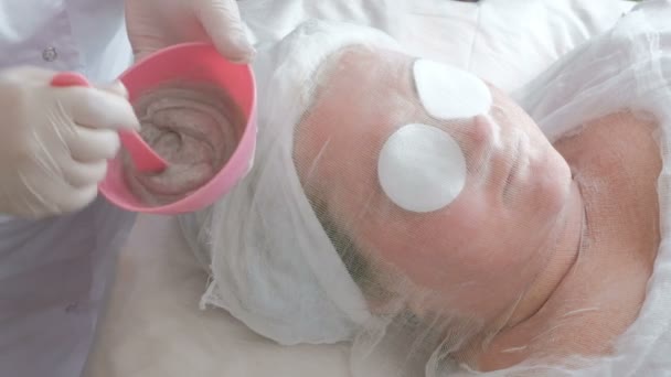 Una chica con gasa en la cara y almohadillas de algodón en los ojos yace en el sofá del salón de belleza. Primer plano de la cara y las manos de una mujer. El esteticista con guantes blancos agita la masa de alginato en una taza de goma rosa . — Vídeo de stock