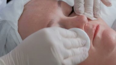 Yüz cilt bakım yordam bir kozmetik merkezinde kapalı gözlü şişman bir kız. Yakın çekim. Yanaklar ve çene bir kadının güzellik uzmanı peçete ile krem kalıntıları kaldırır.