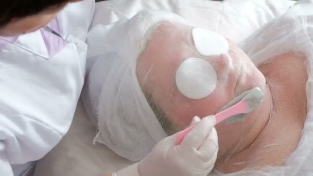 Procédure thérapeutique de nettoyage et de soulèvement de la peau du visage. Le cosmétologue frotte un masque d'alginate de couleur grise sur la joue d'une grosse femme . — Video