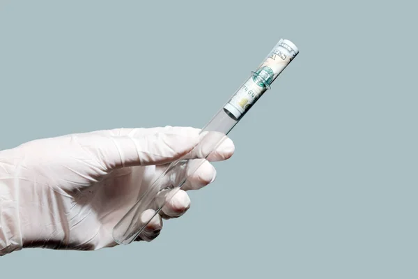 Laboratory test tube with a hundred dollar bill inside in the left male hand. White rubber glove. Concept: money and medicine, corruption and laboratory, science and finance. Isolated gray background.