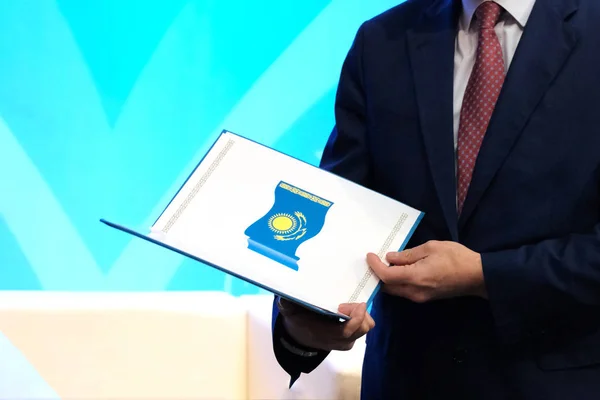 The concept of awarding a civil servant. A man in a dark suit holds a opened folder with the image of the national flag of the Republic of Kazakhstan. Blue background. Copy space.