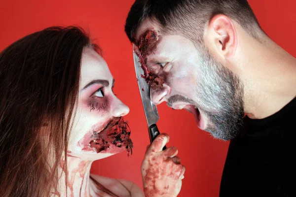 Halloween concept of makeup for All Saints or the Dead. The guy screams at the time when the girl cuts his face with a bloody knife. Horrible cosplay. Close-up. Torn wounds on the face and darned mouth.