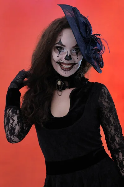 Halloween makeup. Sexy beautiful girl in black dress and cute hat is smiling. Red background. Lace clothes. Body painting on the face. Vertical image. — Stock Photo, Image