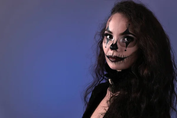Halloween makeup for a party on All Saints Day. Portrait of a beautiful brunette girl with long hair in black clothes on a blue background. Body painting on the face in the style of an evil clown. — Stock Photo, Image