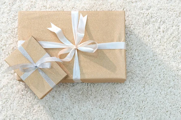 Two boxes with gifts wrapped with golden paper and tied with white ribbon. Soft white carpet. Copy space. Side light.