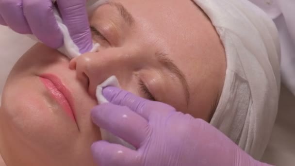 Mulher de meia-idade no centro de cosmetologia. As mãos de um cosmetologista em luvas lilás colocam uma solução de sabão na face feminina. O rejuvenescimento e a limpeza da pele. Close-up de vídeo cru . — Vídeo de Stock